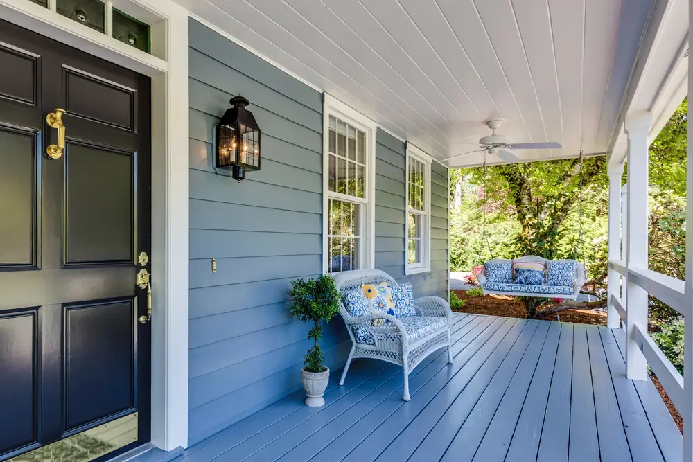 The covered front porch of a blue clapboard rental home with wicker chairs and a porch swing